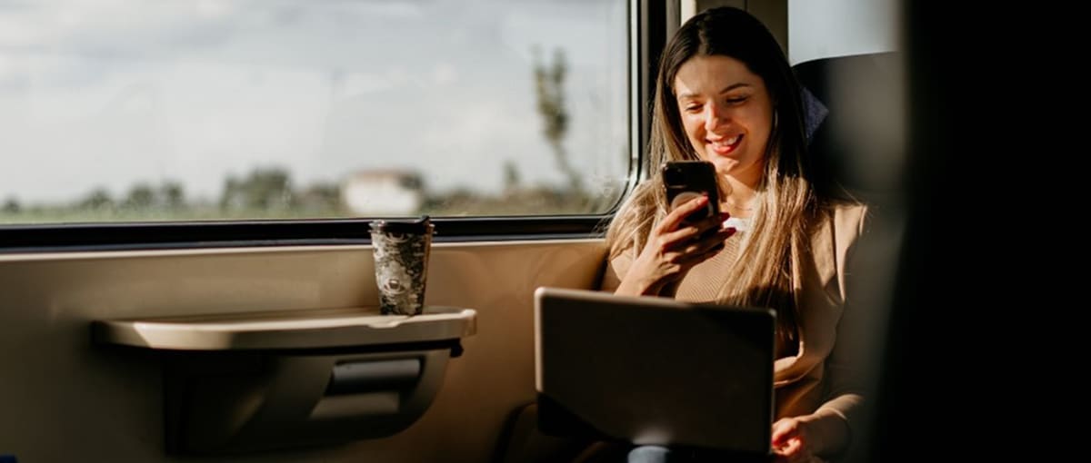 Una persona è seduta al finestrino del treno e usa uno smartphone, mentre ha un laptop sulle ginocchia.