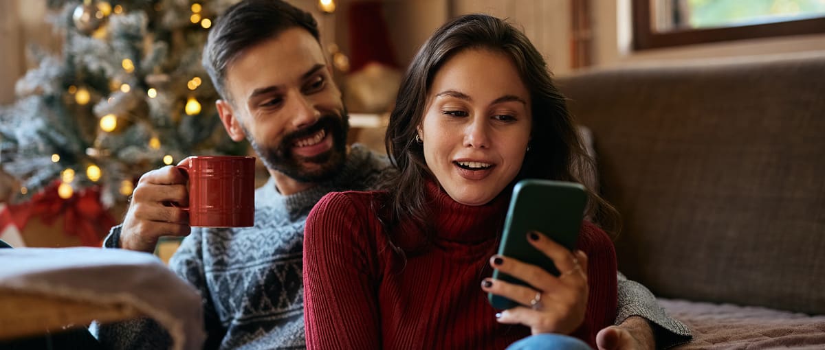 Eine Frau sitzt mit einem Mann und ihrem Handy auf dem Sofa und lächelt.