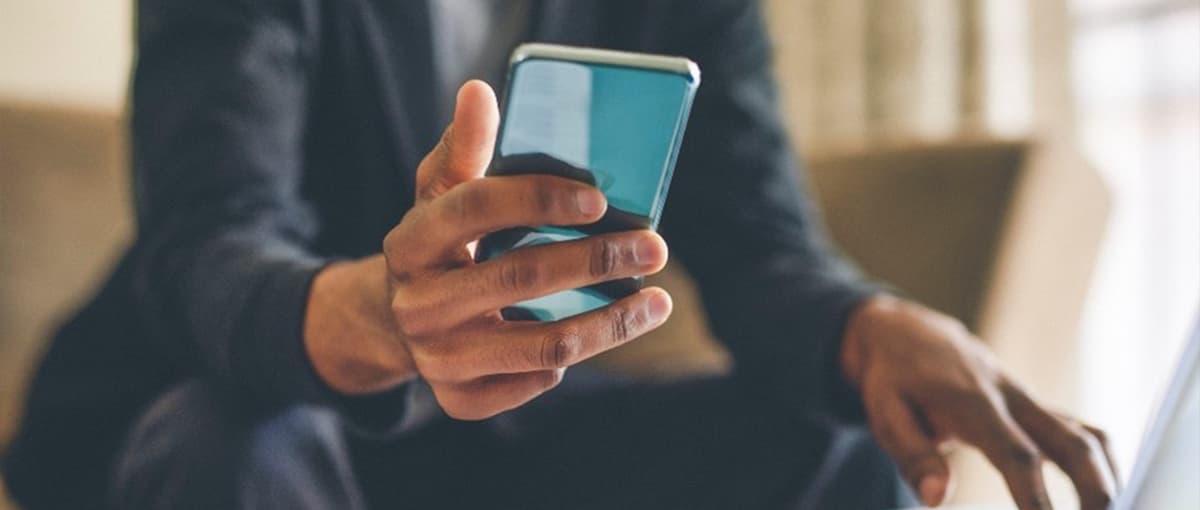 Person hält ein Smartphone in der Hand und bedient gleichzeitig einen Laptop an einem Tisch.