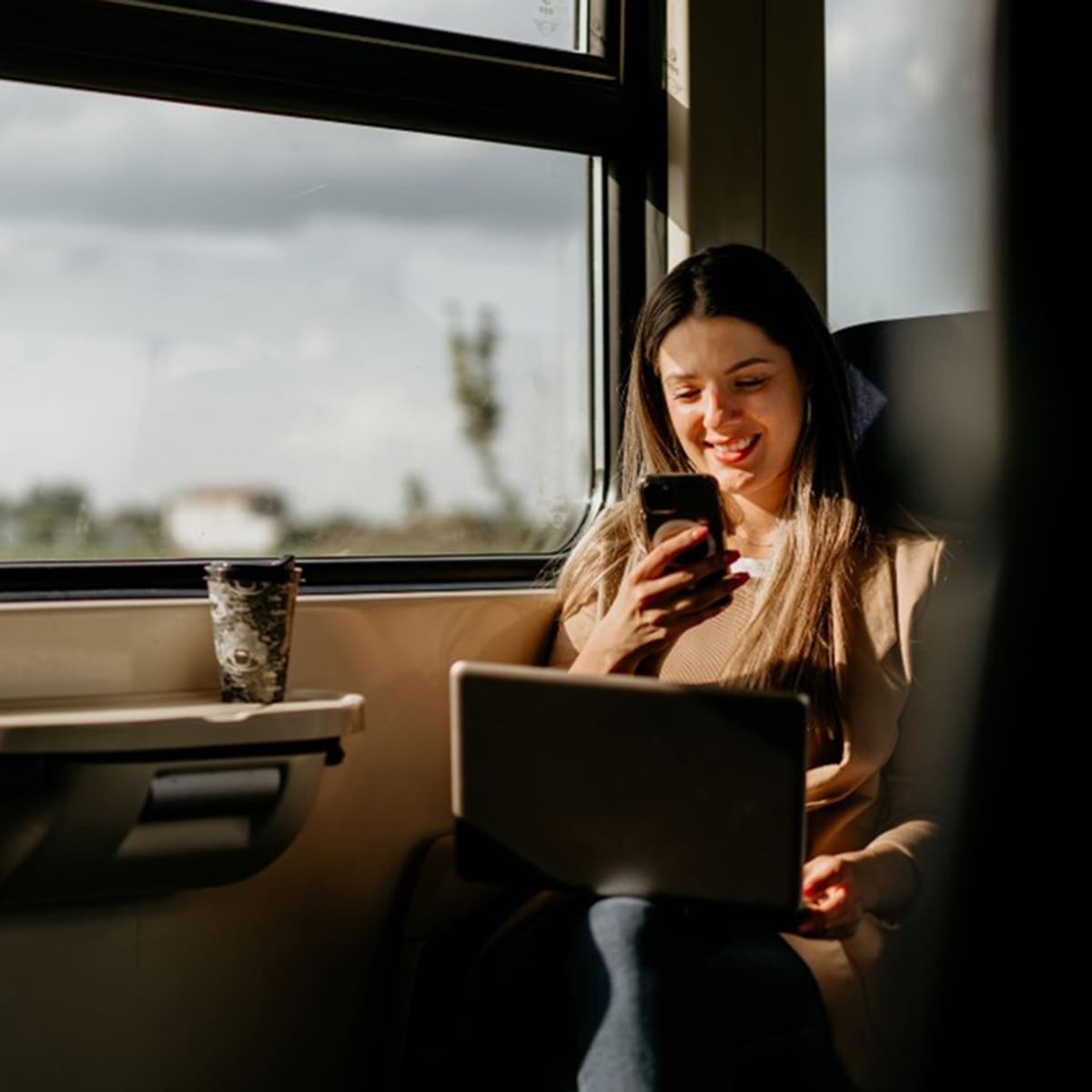 Person sitzt im Zug am Fenster und nutzt ein Smartphone, während ein Laptop auf dem Schoss liegt. 